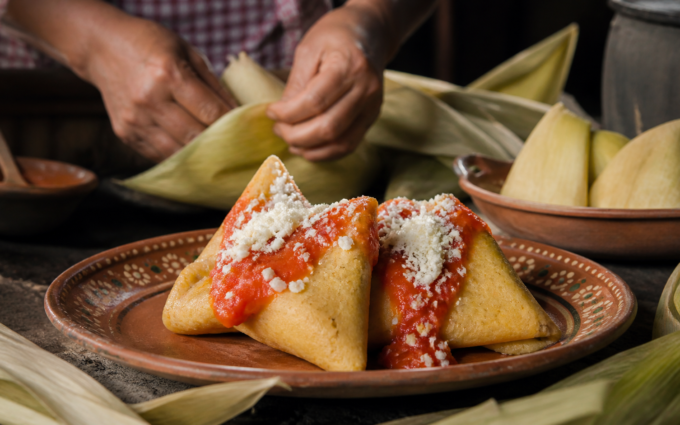 Corundas triangulares de maíz servidas con salsa roja, queso y hojas de caña sobre una mesa de cocina tradicional.